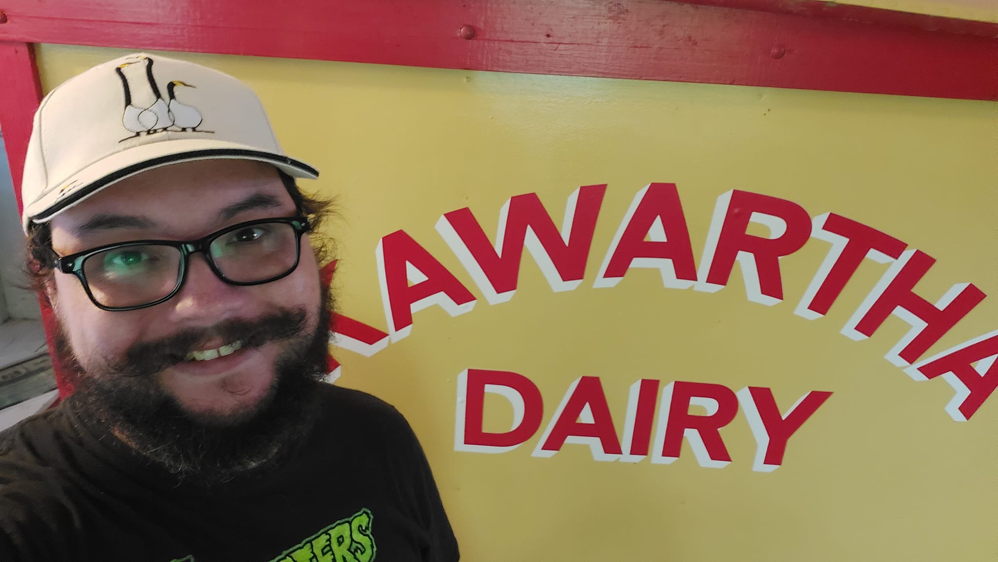 A man with short hair and bushy facial hair stands in front of a the panel of an antique dairy truck (photographer has forgotten if its motorized or horse drawn) with Kawartha Dairy written on it